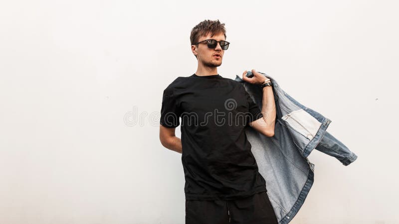 Attractive cool young man hipster puts on a denim jacket. Handsome stylish guy in a vintage T-shirt with trendy sunglasses posing