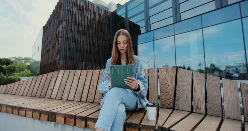 Attractive female sitting on bench and using digital tablet. Summer sunny day. Concept of freelance. Technology concept.