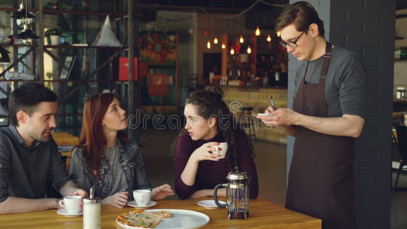 Attractive friends are ordering food and drinks in cafe while friendly smiling waiter in apron is writing order in