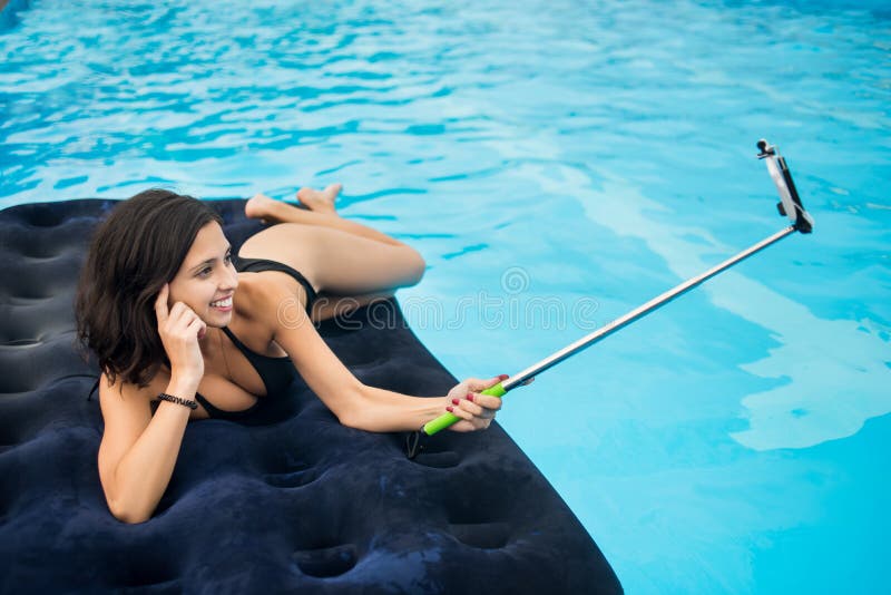 Attractive girl in bikini smiling and makes selfie photo on the phone with selfie stick on a mattress in the pool at the resort