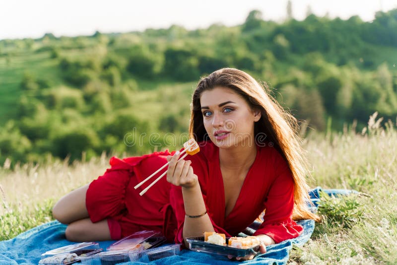 Attractive girl with 3 sushi sets lays on the blue cover on green hills background. Sexy girl in red dress gets sushi