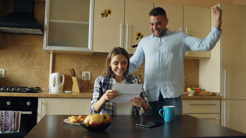 Attractive happy couple recieve good news unfolding letter in the kitchen while have breakfast at home stock photo Attractive happy couple recieve good news unfolding letter in the kitchen while have breakfast at home stock photo