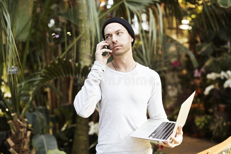 Attractive hipster man talking by mobile phone, using modern laptop computer. Standing in a green park sunny day stock images Attractive hipster man talking by mobile phone, using modern laptop computer. Standing in a green park sunny day stock images