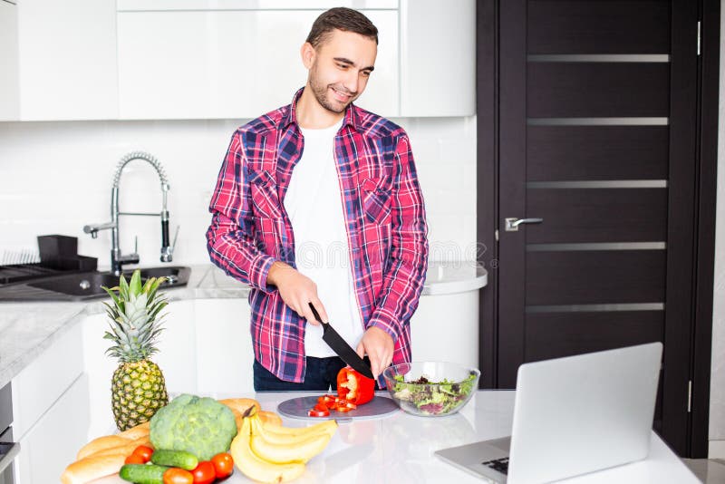 Attractive young man looking on the screen of laptop and cooking on the kitchen stock photography