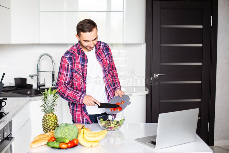 Attractive young man looking on the screen of laptop and cooking on the kitchen royalty free stock photo