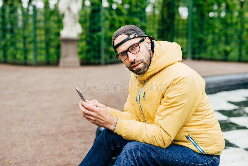 Attractive man with thick beard wearing trendy cap, yellow anorak and glasses sitting outdoors with telephone. Tourist male in cas stock photography Attractive man with thick beard wearing trendy cap, yellow anorak and glasses sitting outdoors with telephone. Tourist male in cas stock photography