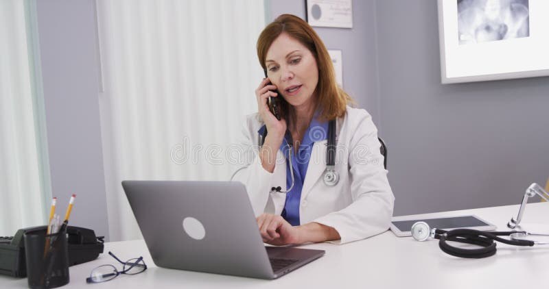 Attractive mid aged doctor using latop computer while talking on smartphone