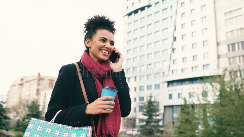 Attractive mixed race business woman talking smartphone and drinking coffee walks in city street with shopping bags stock photo Attractive mixed race business woman talking smartphone and drinking coffee walks in city street with shopping bags stock photo