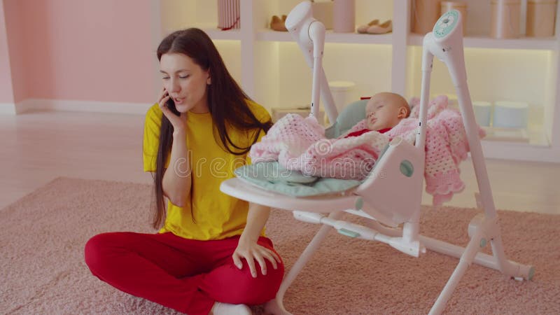 Relaxed mother chatting on phone while infant girl resting in cradle baby swing