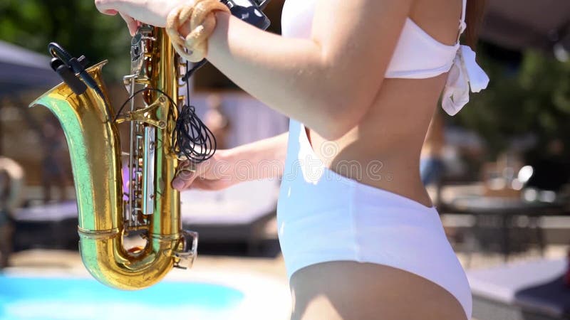 Attractive saxophonist lady is playing on saxophone near pool at beach club. Pretty sax girl musician in hot white