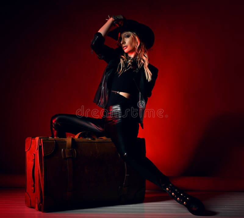 Attractive sexy young blonde woman model in black leather leggins, boots, jacket, hat and accessories sitting on retro