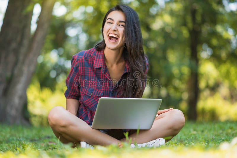 Attractive shot of young Caucasian happy female laughing and wearing plaid shirt, sitting on grass in front of laptop pc, working royalty free stock photography Attractive shot of young Caucasian happy female laughing and wearing plaid shirt, sitting on grass in front of laptop pc, working royalty free stock photography