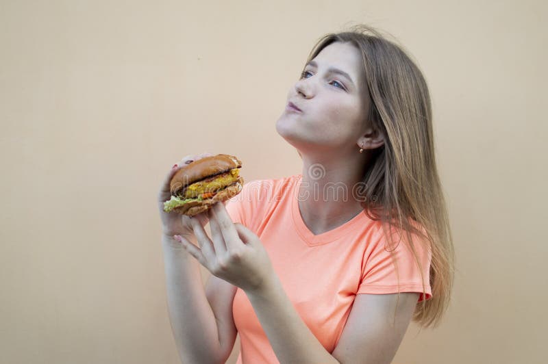 Attractive Teen Girl Stands Against The Orange Wall In An Orange T-shirt And Holds A Big Burger In Her Hand, She Opens Her Mouth