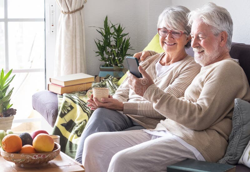 Attractive white-haired senior couple relaxing at home on the sofa looking together at smartphone in video call with family or