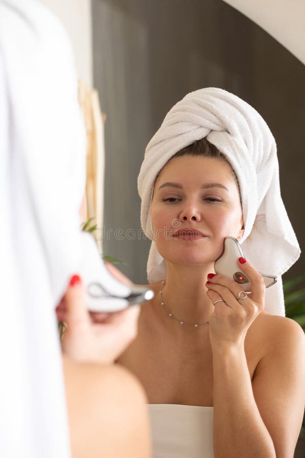 An Attractive Woman Does A Facial Massage With A Microcurrent Massager In Front Of A Bathroom