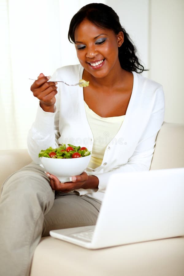 Portrait of an attractive woman eating healthy salad while is sitting on couch in front of her laptop