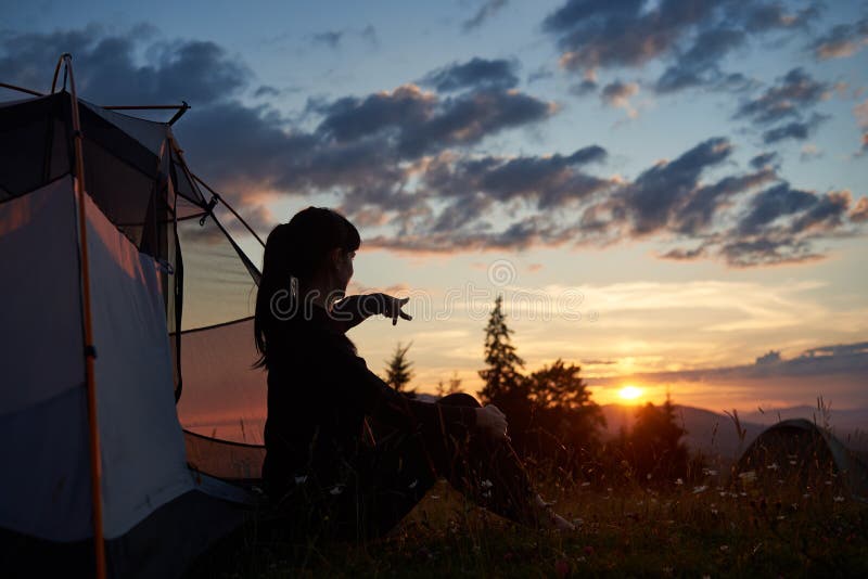 Attractive woman sits in profile near tent on grass with wildflowers and shows her hand in distance