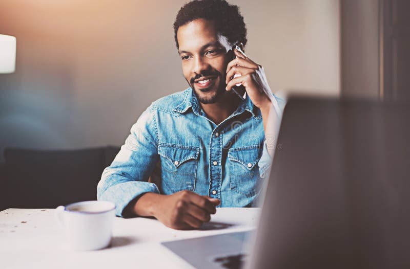 Attractive young African man talking by smartphone while sitting at the wooden table his modern home.Concept of young royalty free stock photos Attractive young African man talking by smartphone while sitting at the wooden table his modern home.Concept of young royalty free stock photos