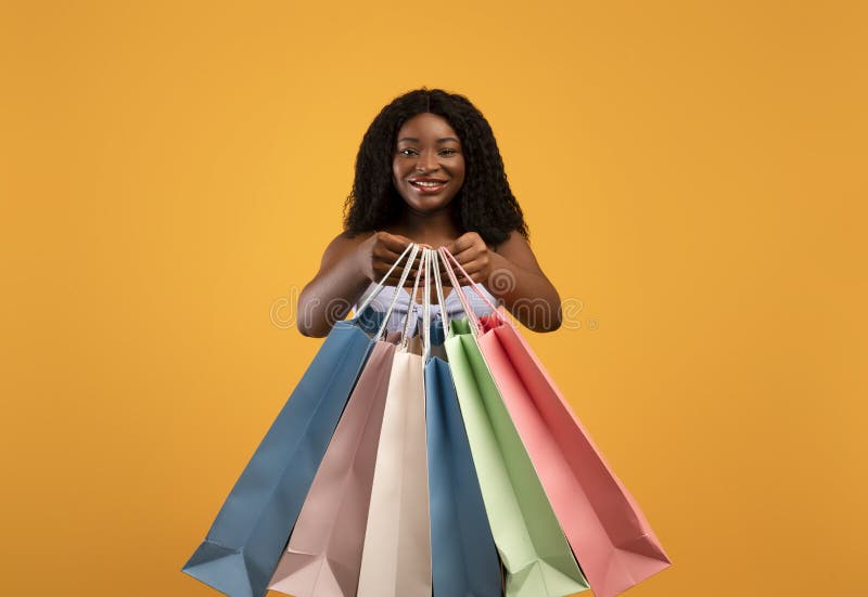 Attractive young black woman holding bright shopping bags, buying goods on sale over orange studio background