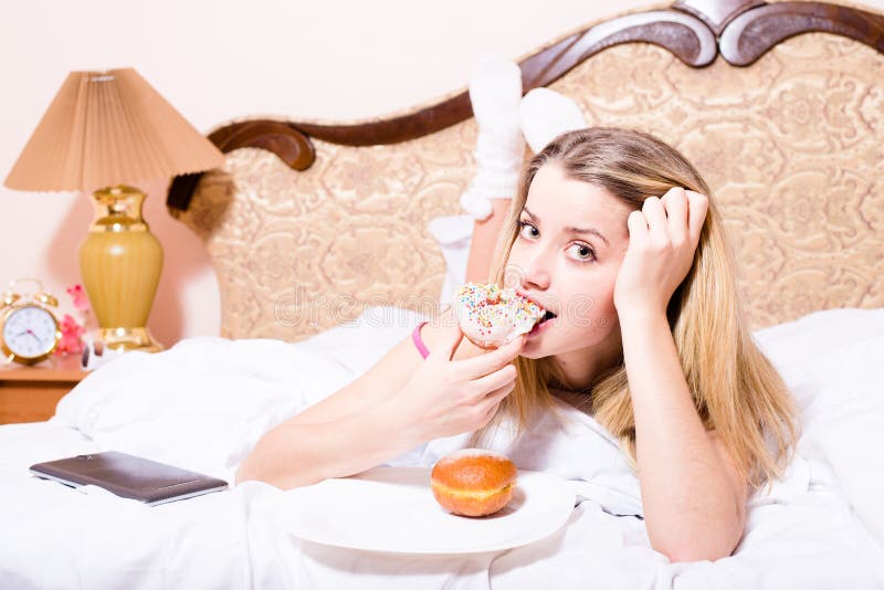 Attractive young blond woman lying in white bed biting color glace donut with tablet pc computer & looking at camera