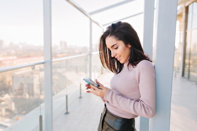 Attractive young brunette woman typing on phone on terrace on big city view background. Sunny morning, relaxing time