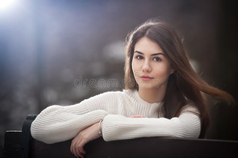 Attractive young Caucasian girl wearing a white blouse sitting on a bench in park. Beautiful brown hair teen girl