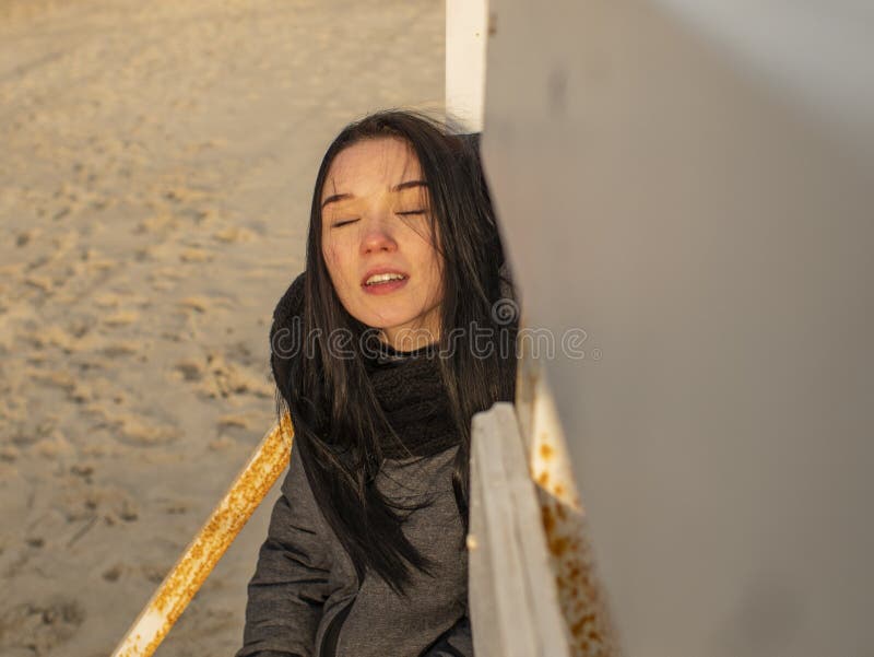 Attractive young girl with disheveled hair sits on a wooden design on the beach in a coat stock photos Attractive young girl with disheveled hair sits on a wooden design on the beach in a coat stock photos