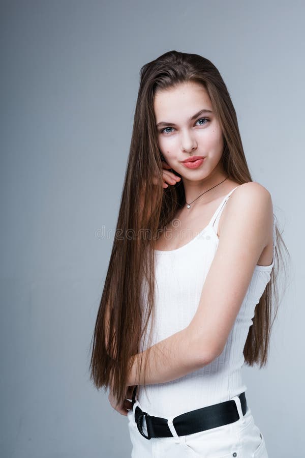 Attractive young girl in a white tank top and trousers with long shiny hair stands sideways and looks at the camera