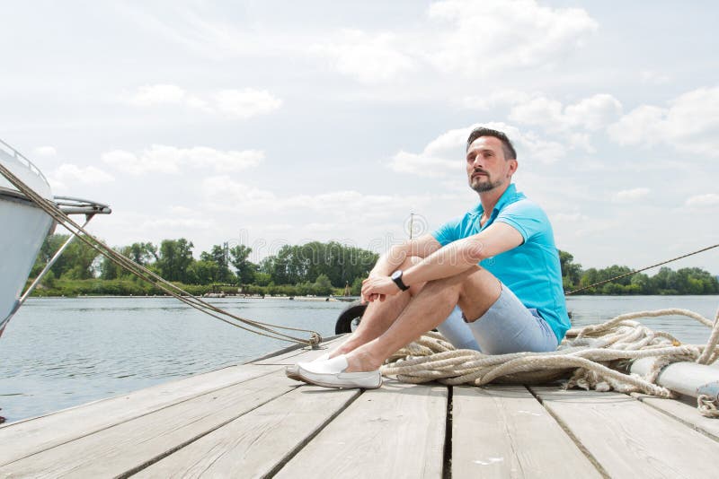 Attractive young guy sits in blue polo on pier near the yacht. smiled tourist handsome man relaxing and enjoying the view on pier royalty free stock photo Attractive young guy sits in blue polo on pier near the yacht. smiled tourist handsome man relaxing and enjoying the view on pier royalty free stock photo