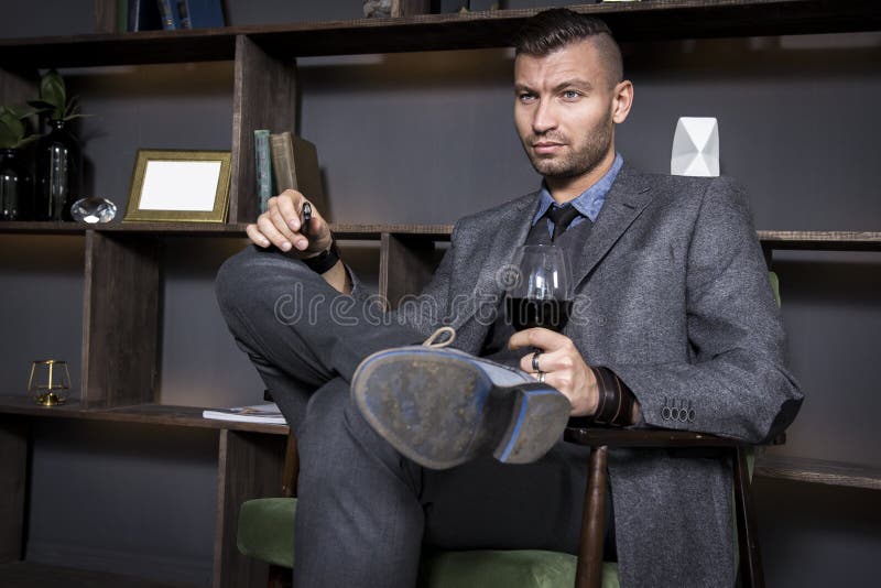Attractive young handsome stylish man in suit sits in chair with glass of red wine. Fashionable elegant man in luxurious interior royalty free stock image Attractive young handsome stylish man in suit sits in chair with glass of red wine. Fashionable elegant man in luxurious interior royalty free stock image