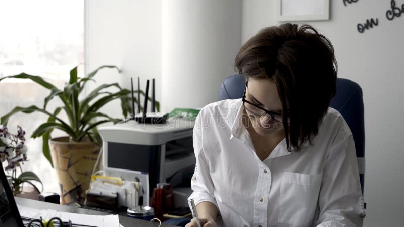 Attractive young office woman in white shirt working on the business papers, signing documents near green plant and royalty free stock photography Attractive young office woman in white shirt working on the business papers, signing documents near green plant and royalty free stock photography