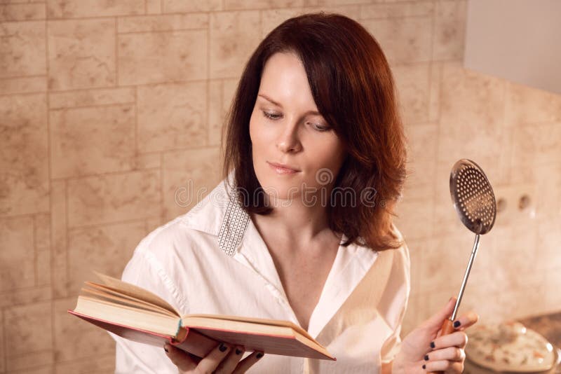 Attractive young redhead woman reading a book while cooking on kitchen