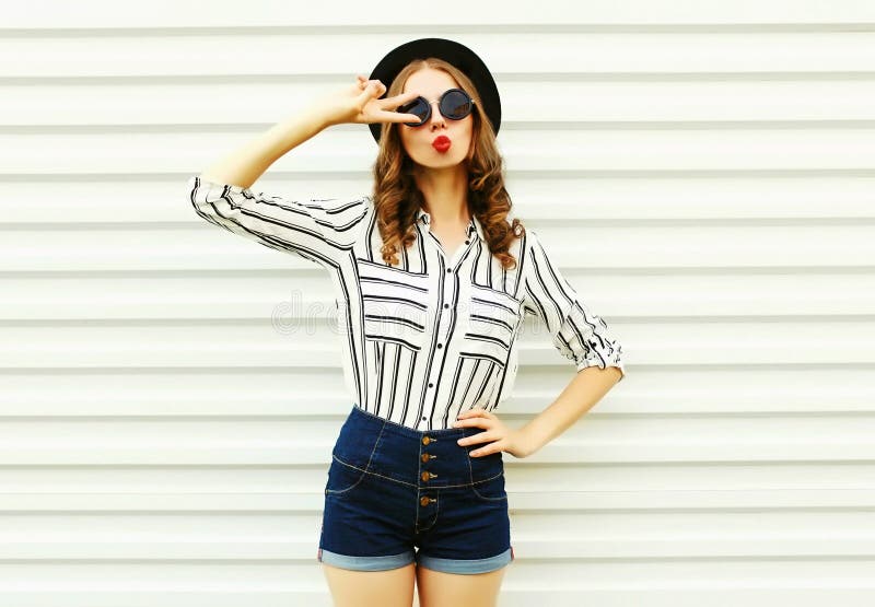 Attractive young woman blowing red lips sending sweet air kiss in black round hat, shorts, white striped shirt on white wall. Background stock image Attractive young woman blowing red lips sending sweet air kiss in black round hat, shorts, white striped shirt on white wall. Background stock image