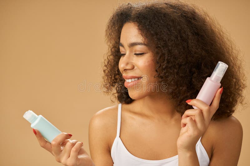 Attractive young woman with curly hair looking cheerful while choosing between two beauty products, posing isolated over