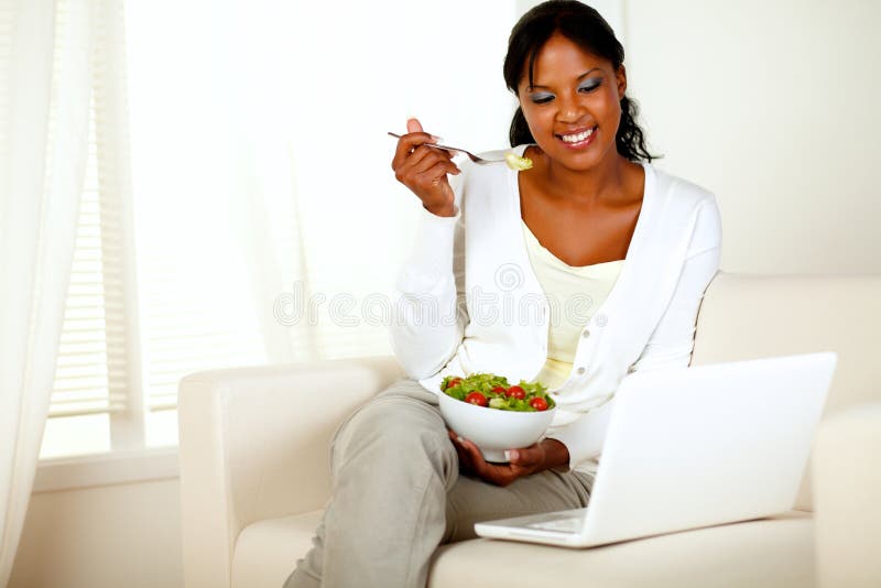 Portrait of an attractive young woman eating healthy salad while is sitting on couch in front of her laptop. with copyspace