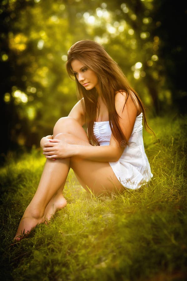 Attractive young woman in white short dress sitting on grass in a sunny summer day. Beautiful girl enjoying the nature