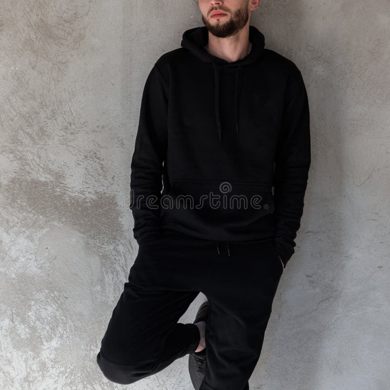 Attraktiv brutal ung man i en trendig svart tröja i svart jeans för tappning med ett stilfullt skägg som poserar på gatan arkivfoto Attraktiv brutal ung man i en trendig svart tröja i svart jeans för tappning med ett stilfullt skägg som poserar på gatan arkivfoto
