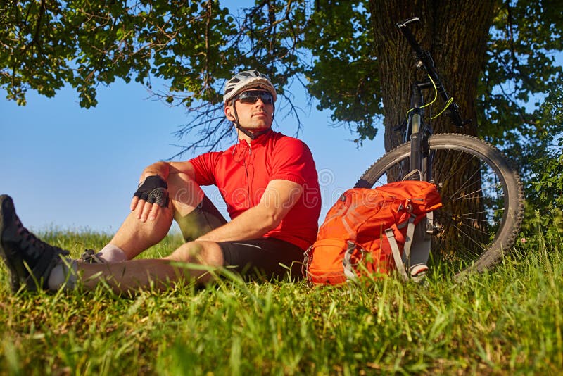Attraktiv cyklist i hjälmsammanträdet på den gröna ängen nära cirkuleringen i bygden royaltyfri fotografi Attraktiv cyklist i hjälmsammanträdet på den gröna ängen nära cirkuleringen i bygden royaltyfri fotografi