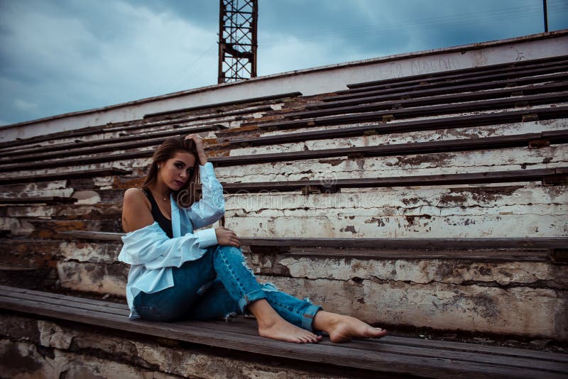 Attraktiv kvinna som sitter med kal fot i stadion Hon bär en skjorta och jeans royaltyfri foto Attraktiv kvinna som sitter med kal fot i stadion Hon bär en skjorta och jeans royaltyfri foto