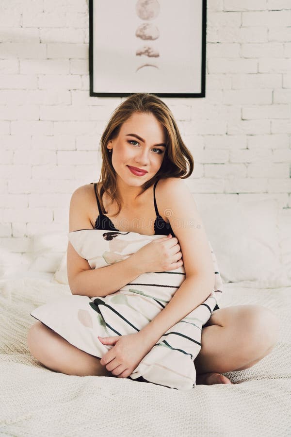 Attraktiv ung kvinna i sexig damunderkläder som sitter i säng med en kudde arkivfoto Attraktiv ung kvinna i sexig damunderkläder som sitter i säng med en kudde arkivfoto