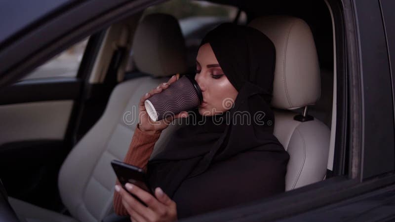 Attraktiv ung kvinna i svart hijab som sitter i sin bil och som fundersamt tittar på sin smartphone-skärm Chatt eller