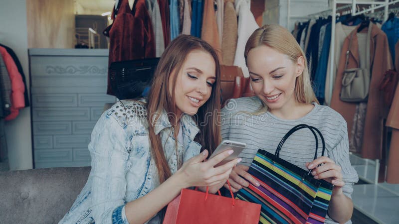 Attraktiva unga damer som sitter i en klädaffär med kaffe- och shoppingväskor som kontrollerar smartphone och pratar royaltyfria foton
