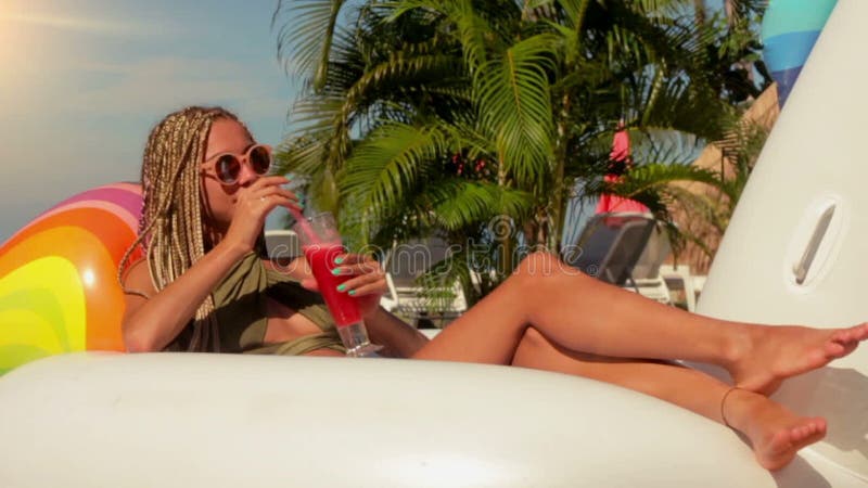 Attraktive Frau mit dem roten Cocktail, das auf aufblasbarem Einhorn im Swimmingpool in der Zeitlupe sitzt