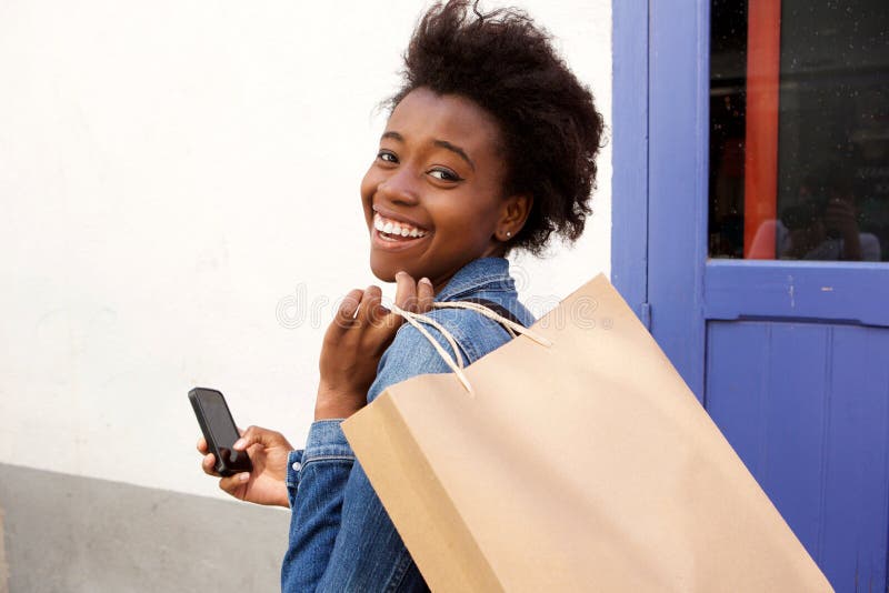 Attraktive junge Afroamerikanerfrau, die mit Telefon und Einkaufstasche lächelt lizenzfreie stockbilder Attraktive junge Afroamerikanerfrau, die mit Telefon und Einkaufstasche lächelt lizenzfreie stockbilder