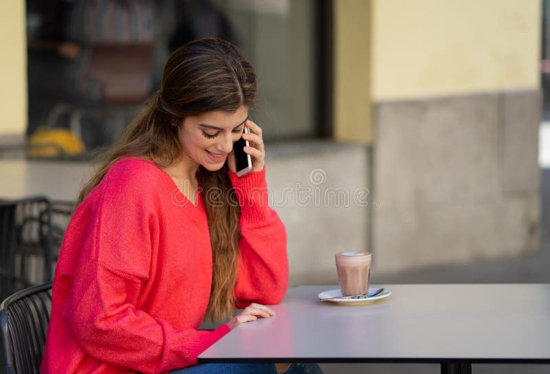 Attraktive junge Frau, die an ihrem intelligenten Handy in einer Kaffeestubeterrasse spricht stockfoto Attraktive junge Frau, die an ihrem intelligenten Handy in einer Kaffeestubeterrasse spricht stockfoto