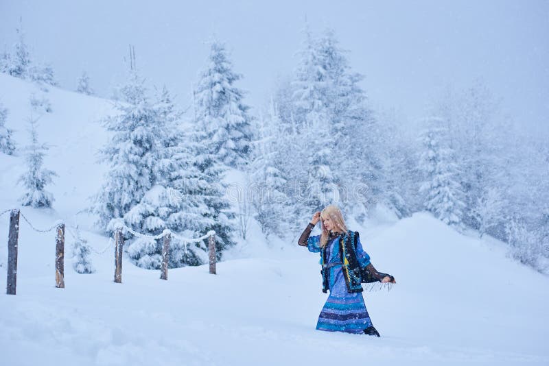 Attractive fashionable slim blond girl in long blue dress, embroidered warm sleeveless fur coat outdoor running in deep clean snow on bright sunny winter day on blurred fir-trees background. Attractive fashionable slim blond girl in long blue dress, embroidered warm sleeveless fur coat outdoor running in deep clean snow on bright sunny winter day on blurred fir-trees background.