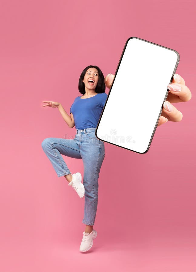 Aufgeregt junge asiatische Dame springt auf und präsentiert Smartphone mit leerem Bildschirm über rosa Hintergrundmockup stockfotos
