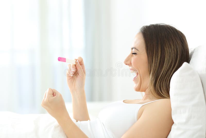 Aufgeregte Frau, die einen positiven Schwangerschaftstest im Bett überprüft