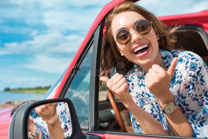aufgeregte junge Frau in der Sonnenbrille, die im Auto sitzt und sich Daumen zeigt lizenzfreies stockbild aufgeregte junge Frau in der Sonnenbrille, die im Auto sitzt und sich Daumen zeigt lizenzfreies stockbild