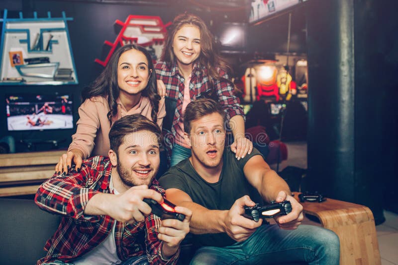 Aufgeregte junge Männer sitzen auf Sofa, wenn sie Raum spielen Sie halten Steuerknüppel und Spielspiel Junge Frauen sitzen hinten stockbild Aufgeregte junge Männer sitzen auf Sofa, wenn sie Raum spielen Sie halten Steuerknüppel und Spielspiel Junge Frauen sitzen hinten stockbild
